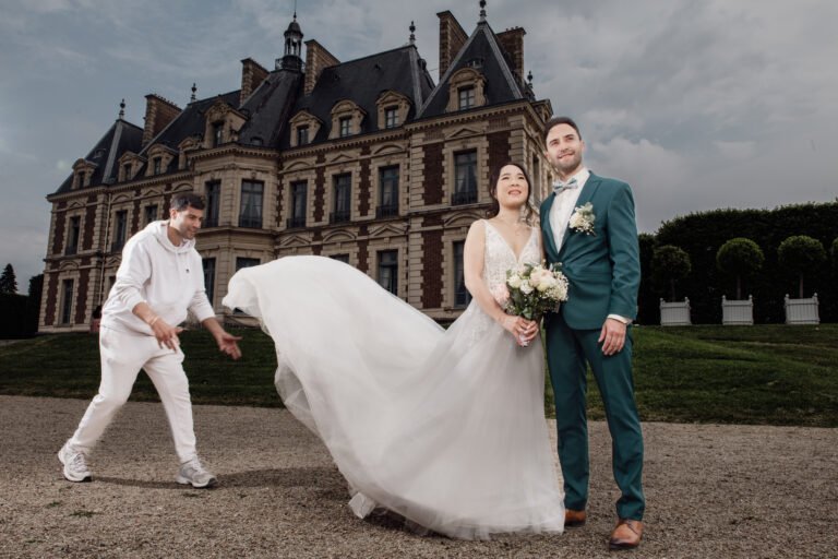 Vidéaste de mariage en blanc secouant la robe de mariée pour un couple devant un château, lumière parfaite, style cinématographique.